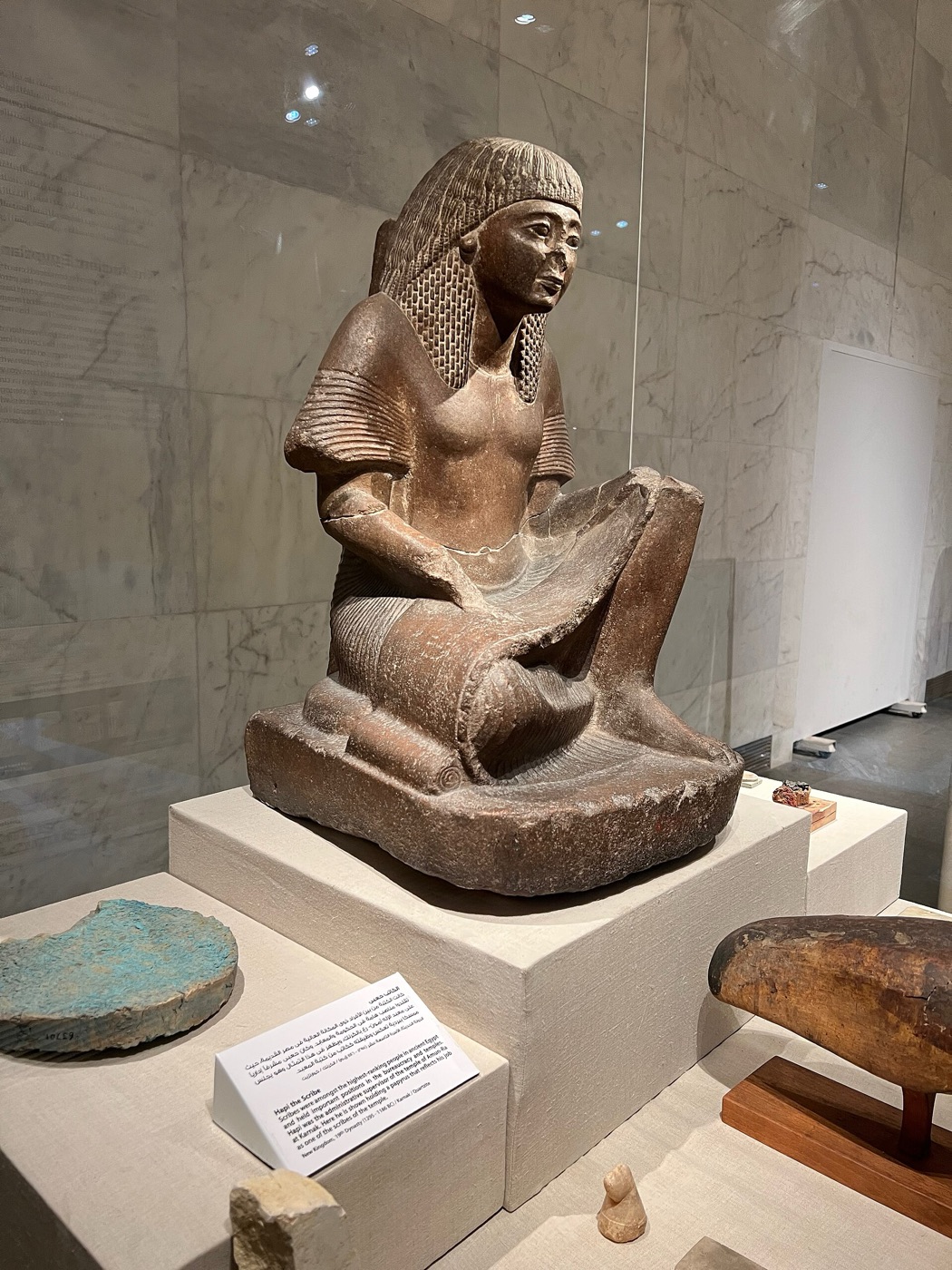 Seated stone statue of Hapi the scribe with a papyrus open across his lap, in a museum case